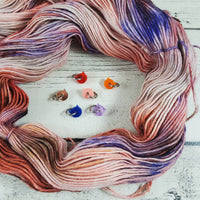 Tiny cat head stitch markers surrounded by a hand-dyed skein of yarn on a white wood background