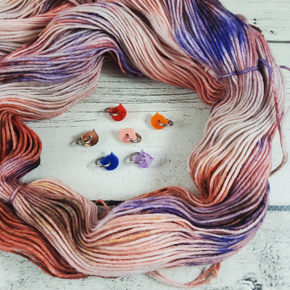 Tiny cat head stitch markers surrounded by a hand-dyed skein of yarn on a white wood background