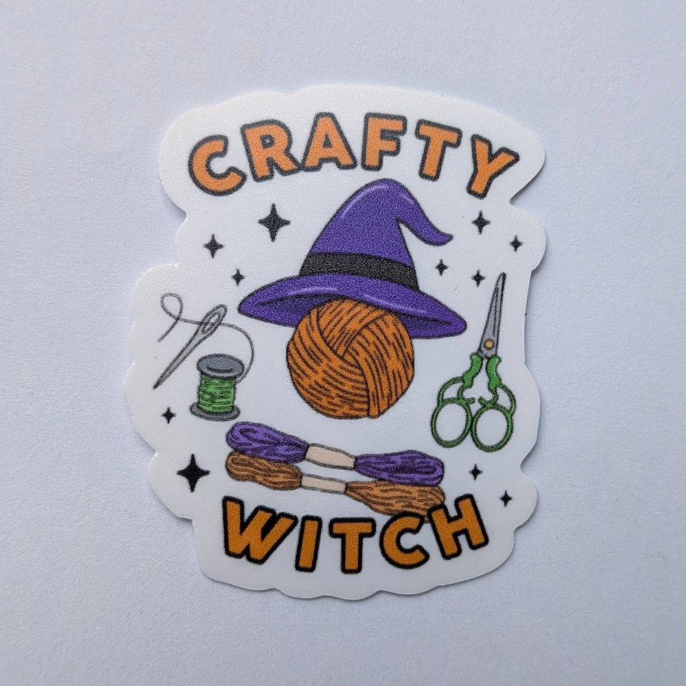 Sticker with 'Crafty Witch' text, yarn ball, witch hat, and crafting tools on a light gray background