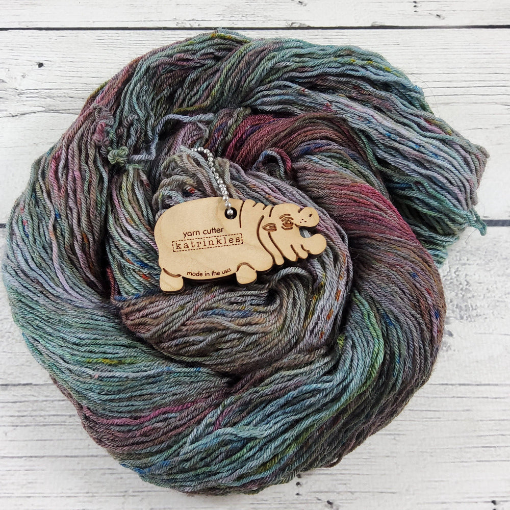 Wood baby hippo yarn cutter on a blue hand-dyed skein of yarn on a white wood background