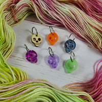 Skull stitch markers surrounded by a green and pink hand-dyed yarn skein on a white wood background
