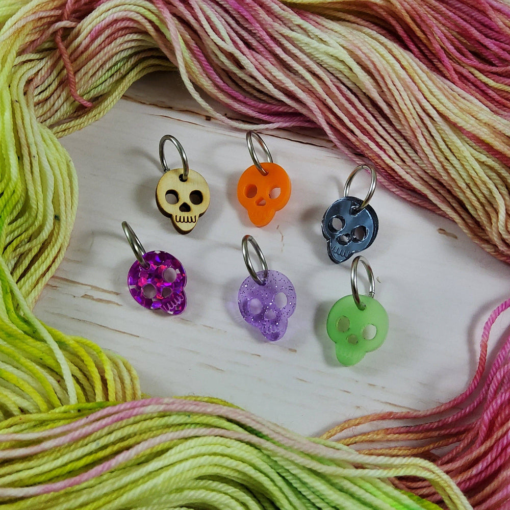 Skull stitch markers surrounded by a green and pink hand-dyed yarn skein on a white wood background