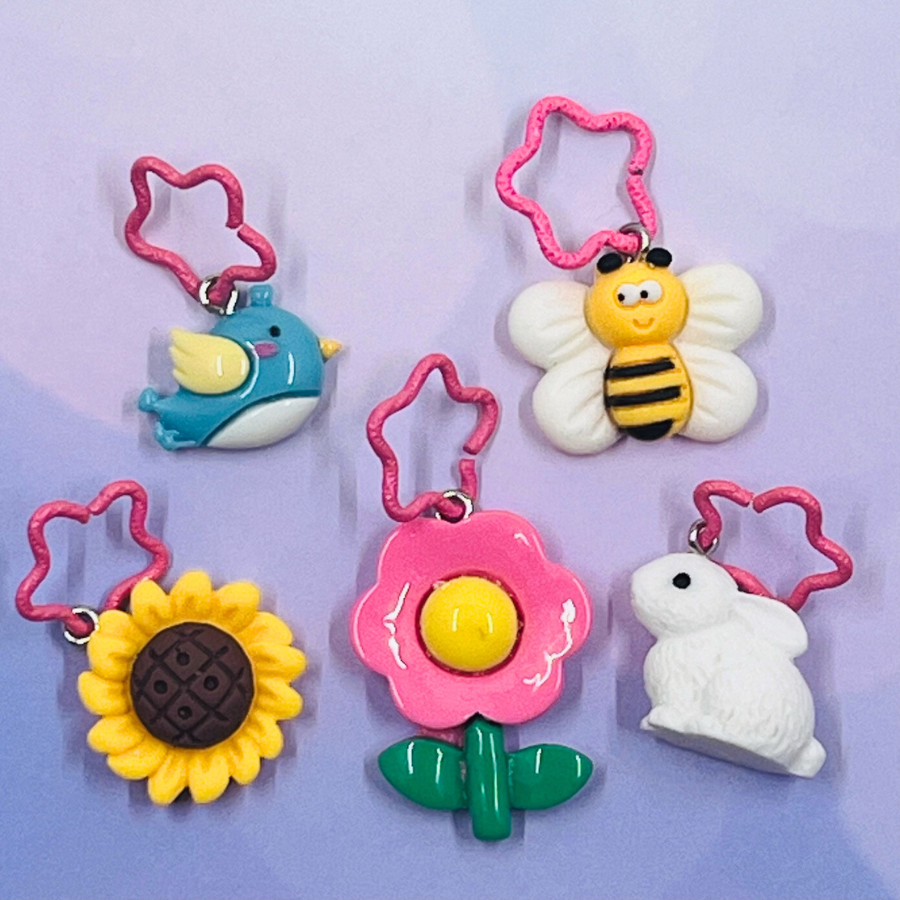 Plastic stitch markers in spring themed shapes: bird, bee, sunflower, pink daisy, and bunny. Each marker is attached to a pink, star-shaped ring.