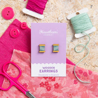 Wooden earrings on a card with pink fabric, buttons, and scissors on a wooden surface