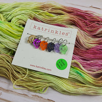 Multicolored skull stitch markers on white "Katrinkles" packaging on top of a green and pink hand-dyed yarn skein with a white wood background