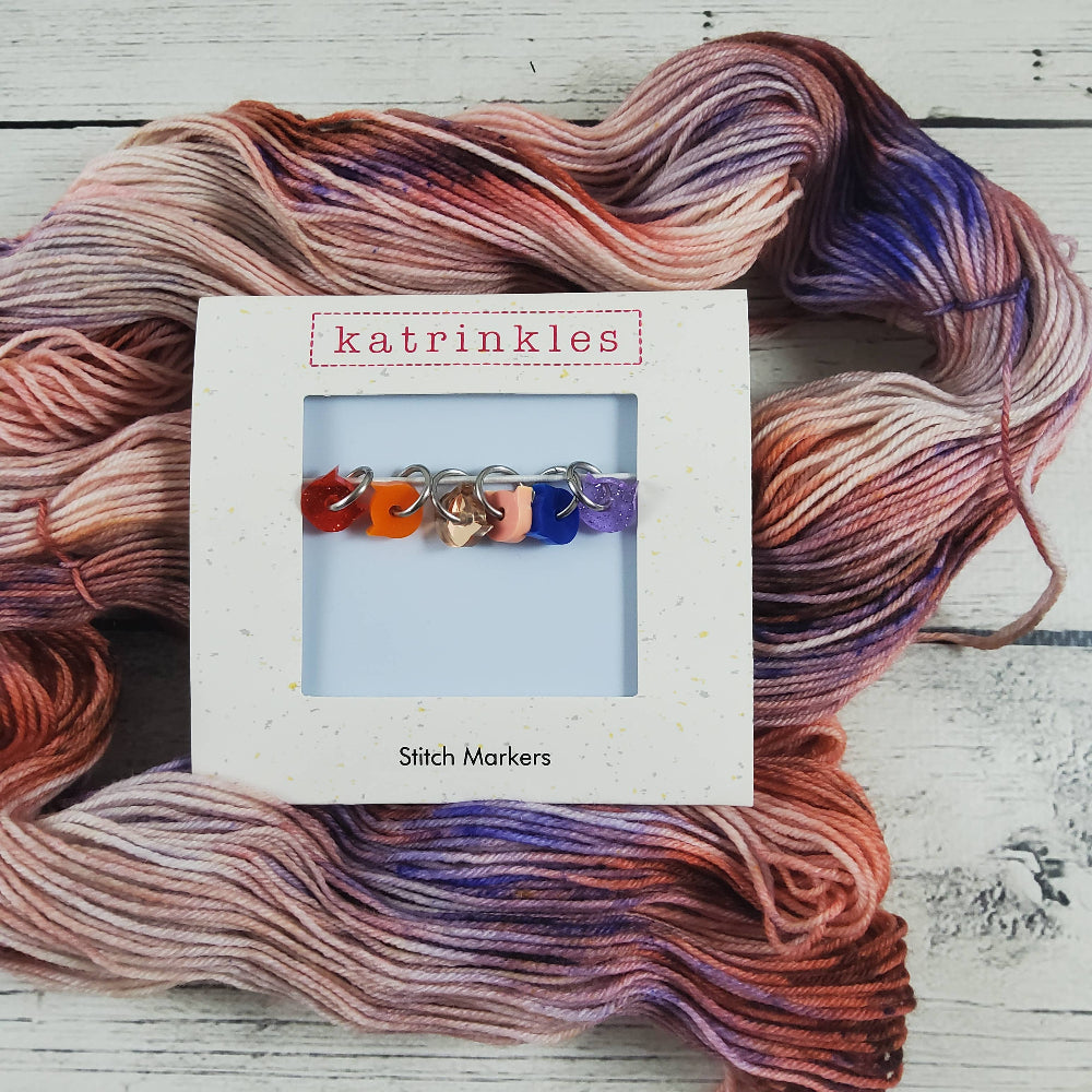 Tiny cat head stitch markers in white packaging titled "Katrinkles Stitch Markers" atop a hand-dyed skein of yarn with a white wood background