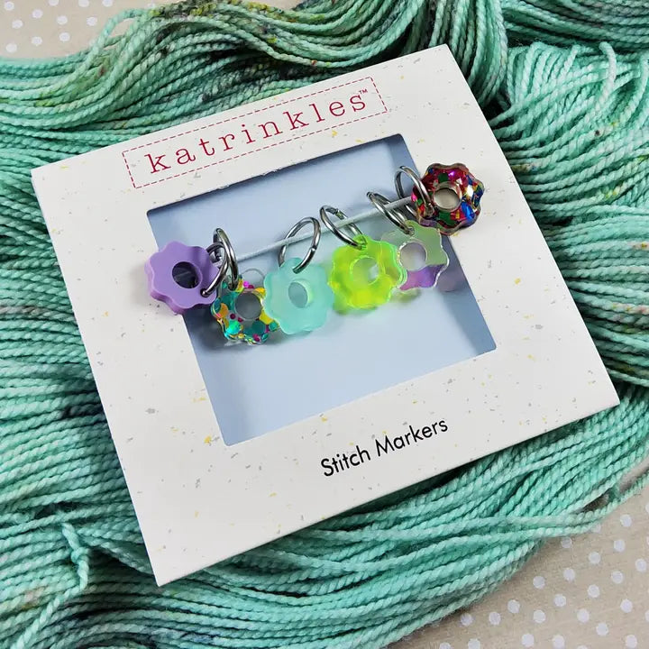acrylic flower stitch markers in different colors and glitters on top of water green yarn.