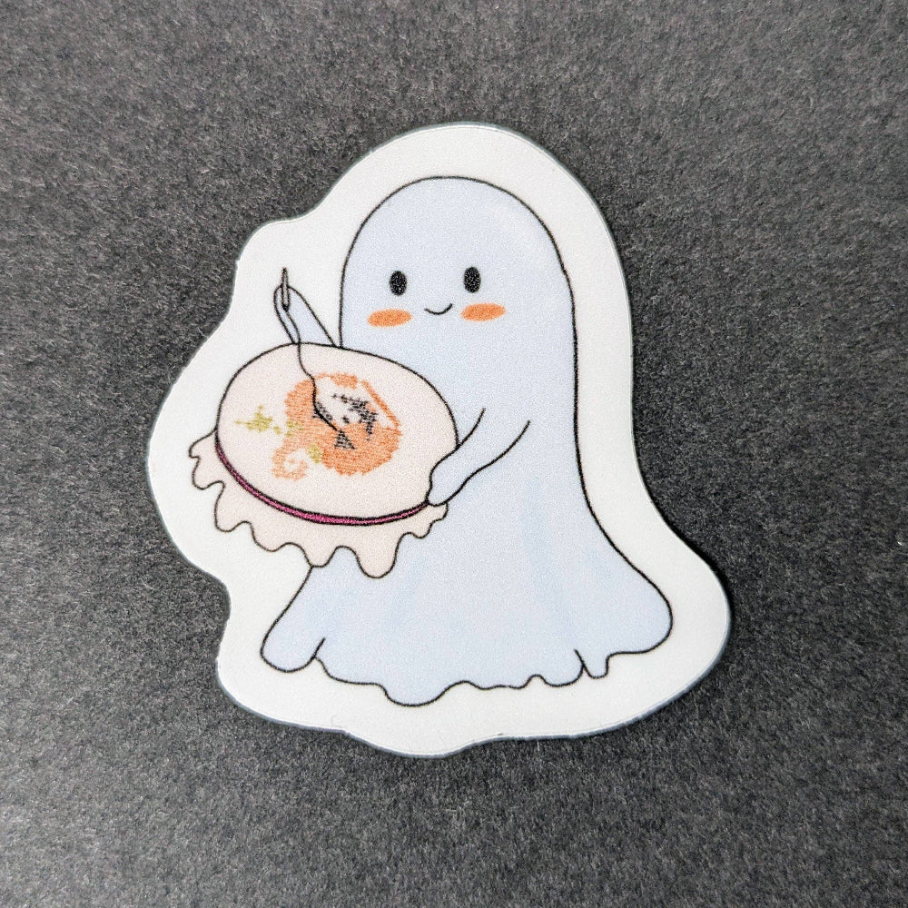 Sticker of a ghost holding an embroidery hoop and needle on a gray background