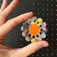 Colorful knitting needle flower gauge held by a hand against a black pegboard background