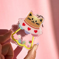 Cat-shaped keychain with colorful design held by a hand against a pink background