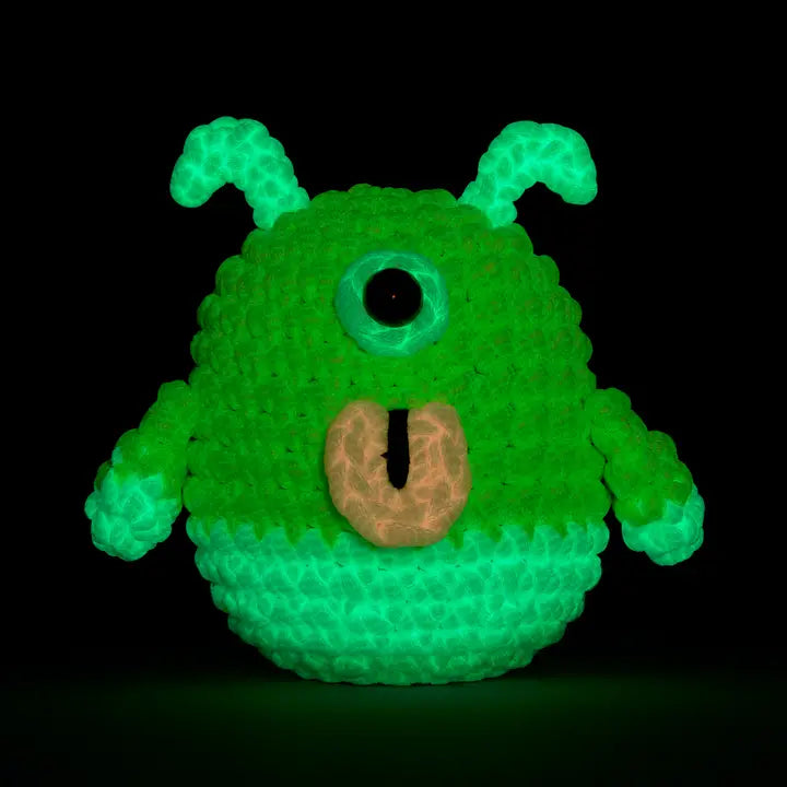 Green crocheted alien with a single eye glowing in the dark on a black background