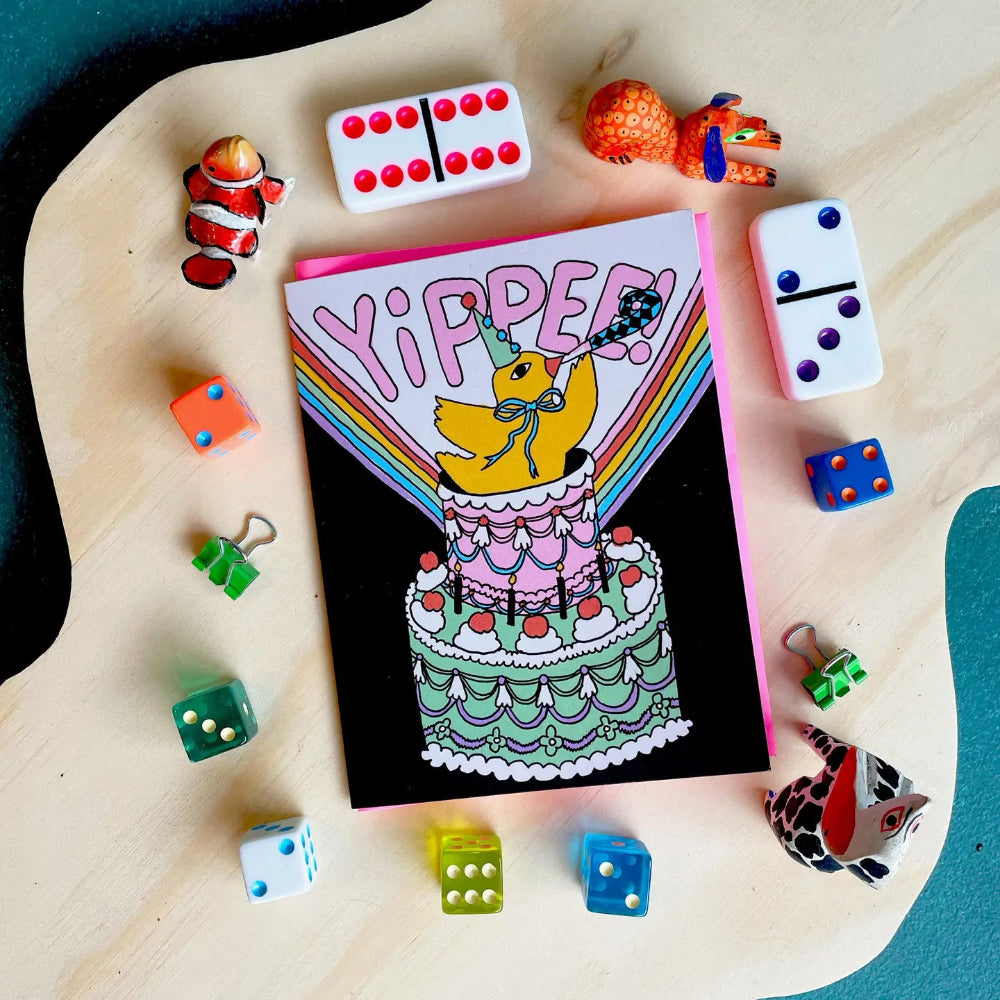 Greeting card with a duck on a cake and 'Yippee!' text, surrounded by colorful dice and game pieces on a wooden surface.