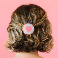 Person with wavy hair wearing a hair clip with a pink heart design on a pink background