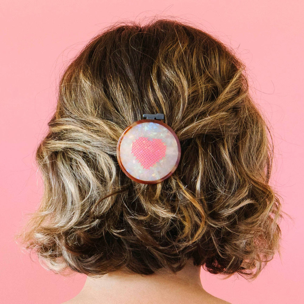 Person with wavy hair wearing a hair clip with a pink heart design on a pink background