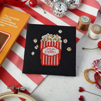 Cross-stitched popcorn design on black fabric with cross stitch supplies around