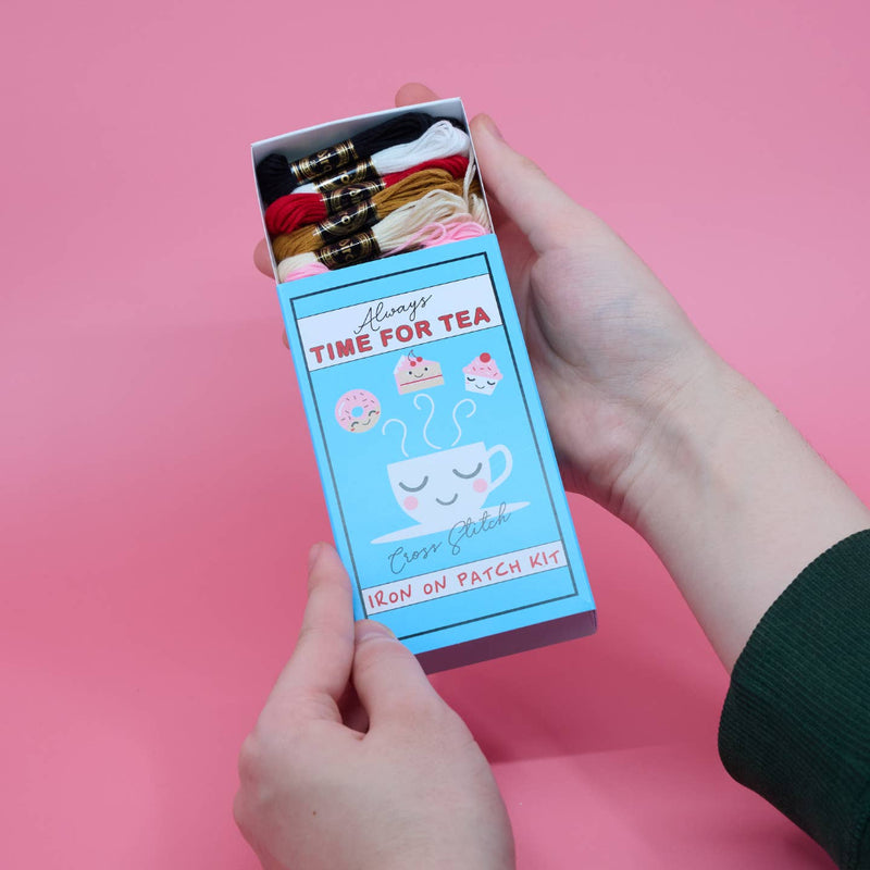 Hand holding a box of 'Always Time for Tea' iron on patch kit against a pink background