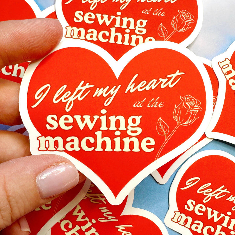 Red heart-shaped sticker with 'I left my heart at the sewing machine' text held by a hand against a blue background.