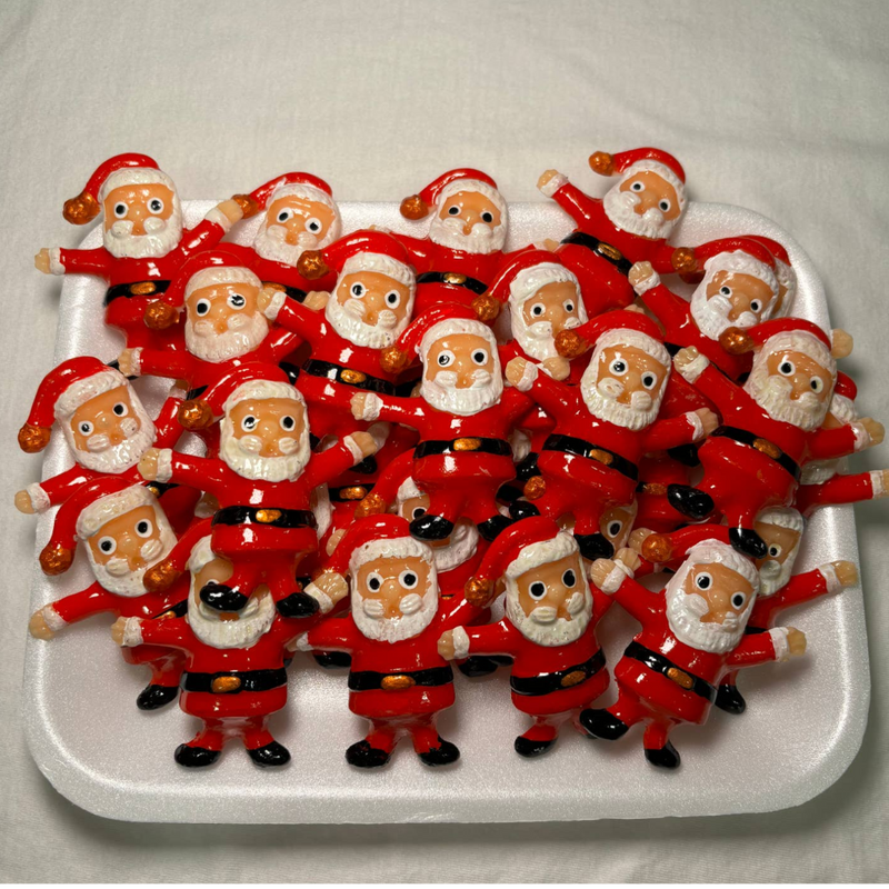 White tray full of clay Santa magnets. Each Santa magnet has a red body, black belt with gold buckle, white beard, and traditional red Santa hat