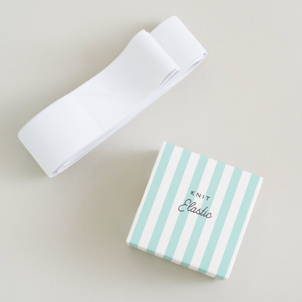 A white elastic band is shown next to its packaging box, which features vertical mint green and white stripes with the brand name 'KNIT Elastic' printed on top, set against a neutral light gray background.