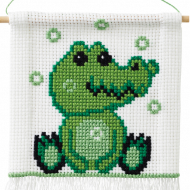 Green crocodile cross stitch on white background with wooden rod on top and white fringe on the bottom.