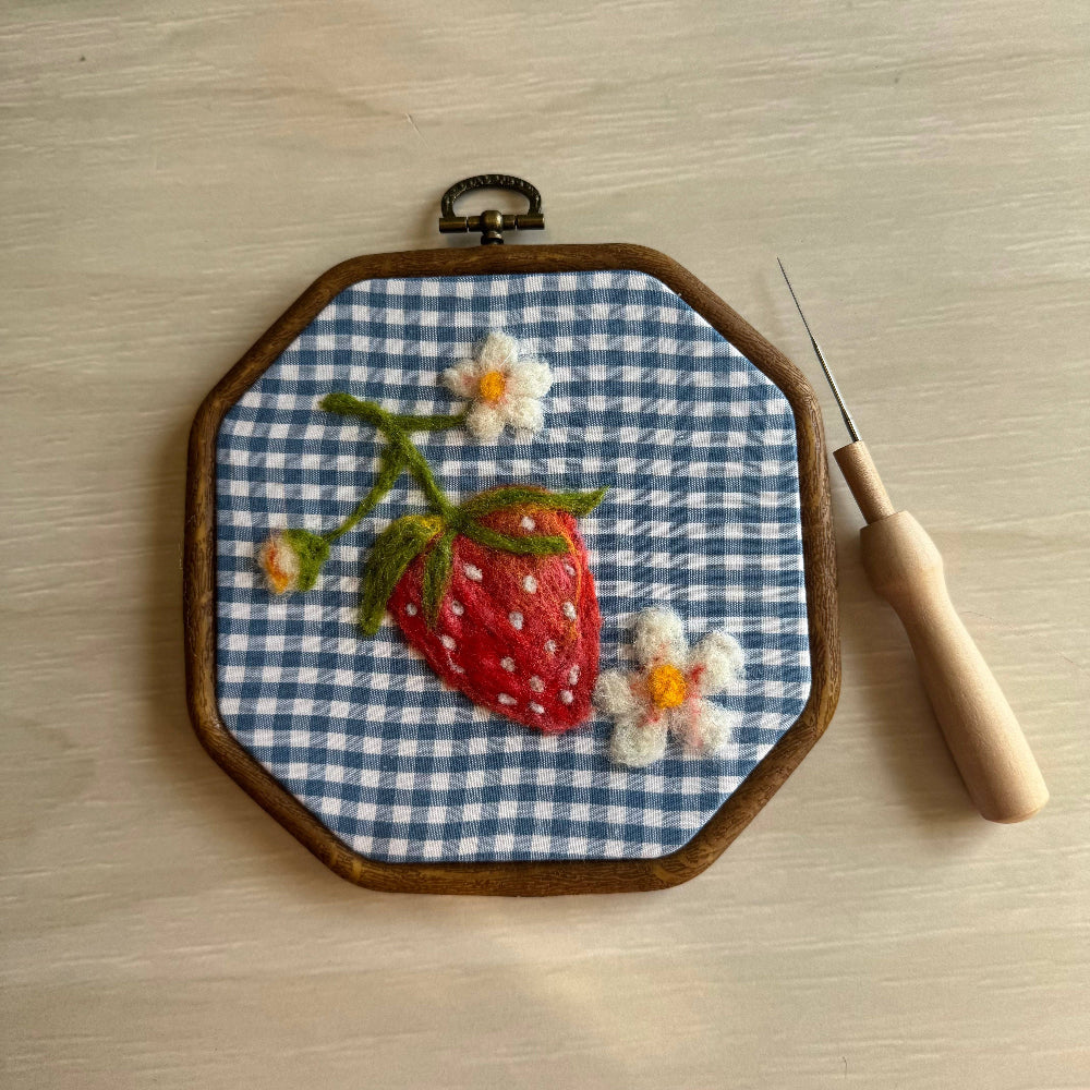 Embroidery hoop with felted strawberry and flowers on blue checkered fabric, accompanied by a needle and wooden handle.