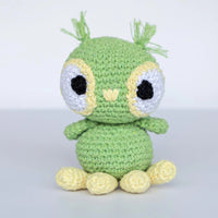 A cute crocheted amigurumi toy with a light green body, large white eyes with black centers, yellow facial details, and four yellow feet, set against a plain light gray background.