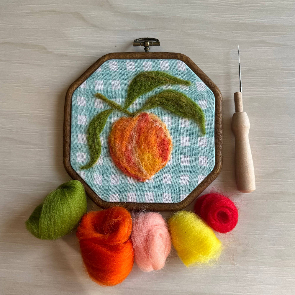 Embroidery hoop with a peach design on checkered fabric, surrounded by colorful yarn balls and a needle.