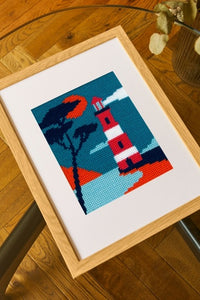 Framed cross-stitched picture of a lighthouse on a wooden table