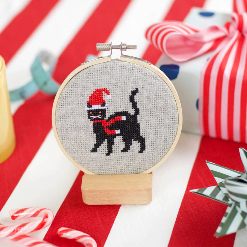 Cross-stitched hoop with a black cat wearing a Santa hat on a red and white striped background.
