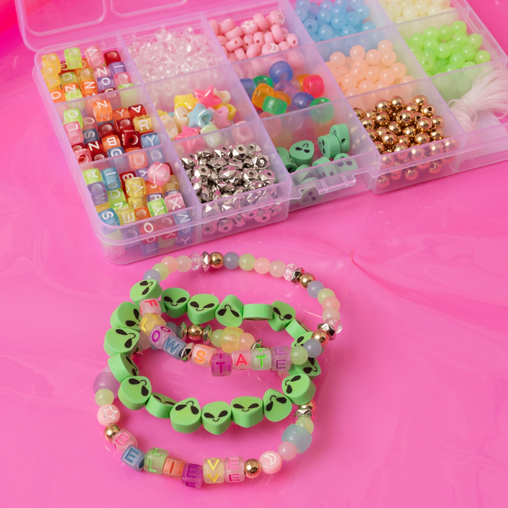 beaded bracelets with alien and letter beads. Theres a box of colorful beads in the back on a pink background.