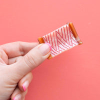 Hand holding a sewing spool shaped hair clip with a pattern against a pink background