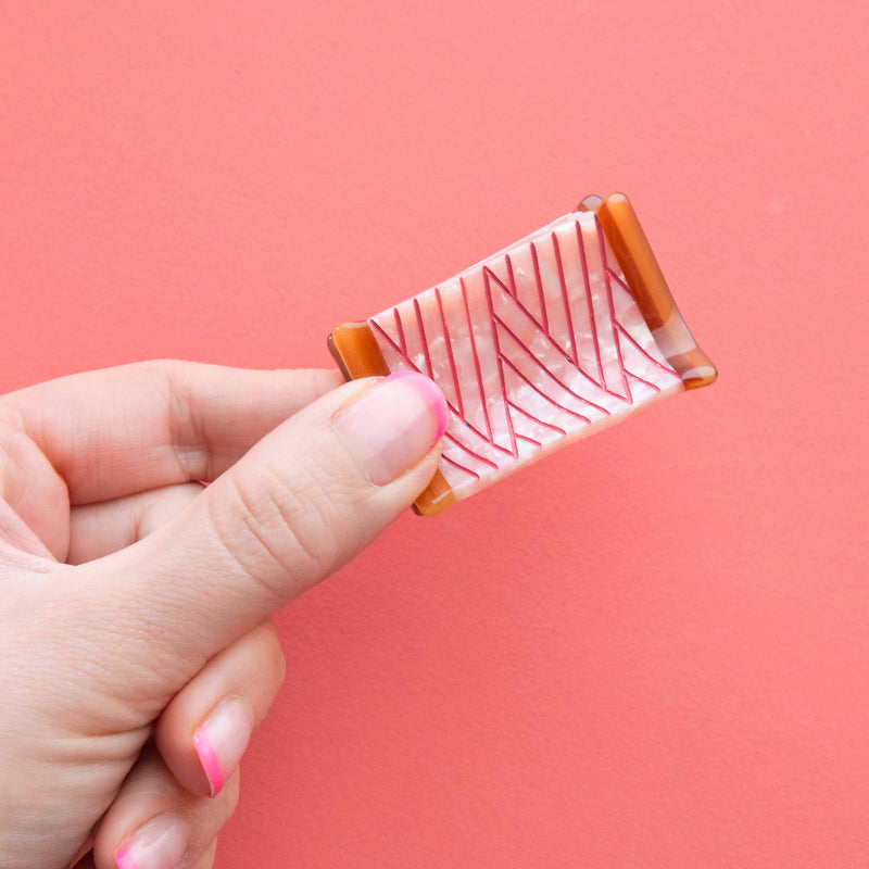 Hand holding a sewing spool shaped hair clip with a pattern against a pink background