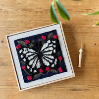 Embroidery of a butterfly and flowers in a box on a wooden surface with a needle next to it.