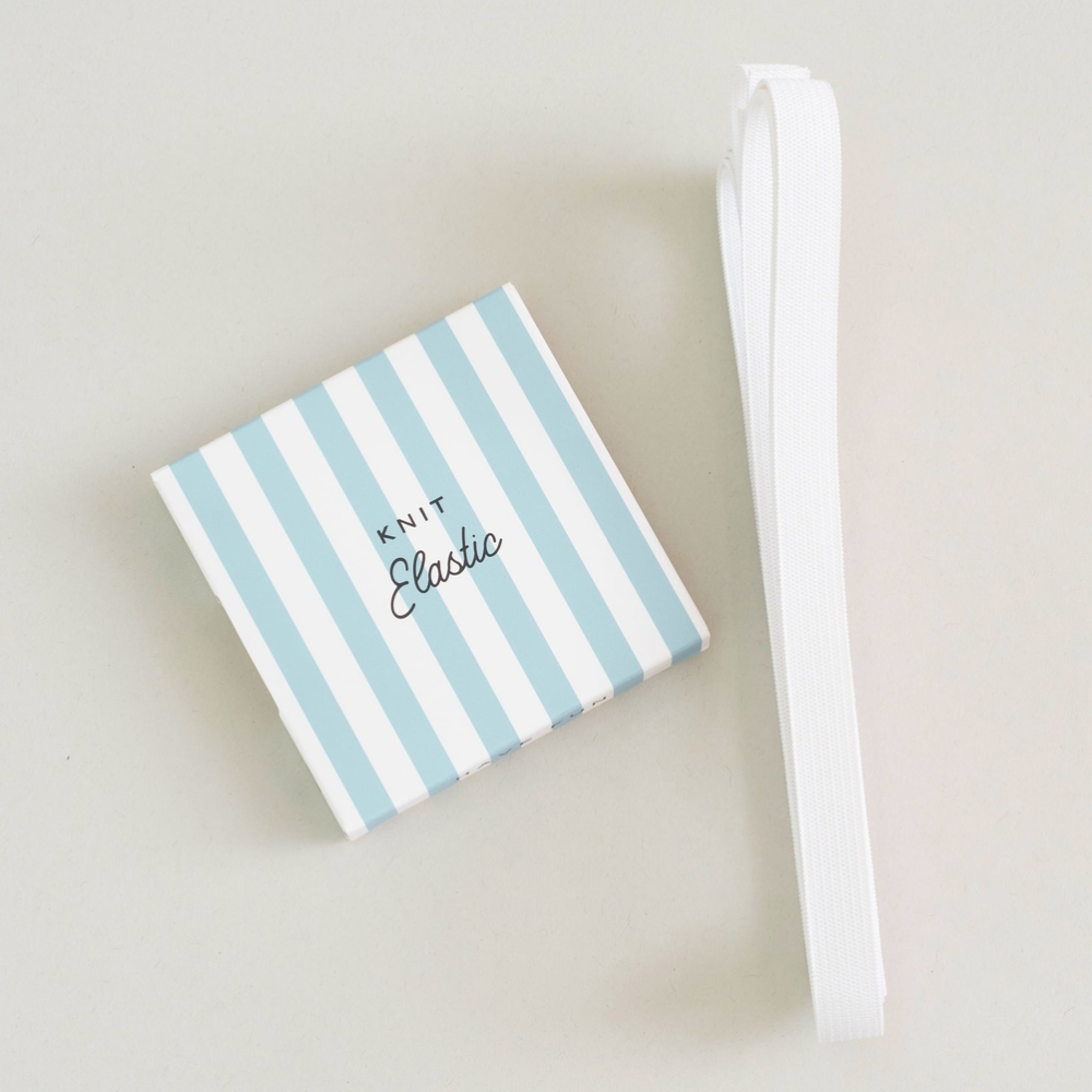 A small square box with light blue and white vertical stripes sits on a light beige surface next to a rolled-up white elastic band. The box is printed with the words 'KNIT Elastic'.