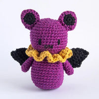 A purple crocheted plush toy with black wings, a yellow ruffled collar, and black circular details on its ears, photographed against a plain white background.