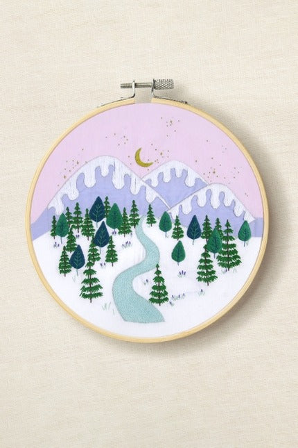 Embroidery hoop with a winter landscape design featuring trees, mountains, and a river.
