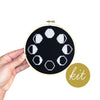 Embroidery hoop with black fabric featuring white phases of the moon, held by a hand. 'Kit' text on a gold circle.