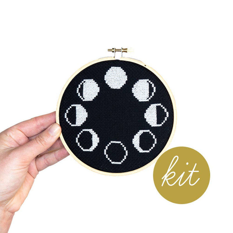 Embroidery hoop with black fabric featuring white phases of the moon, held by a hand. 'Kit' text on a gold circle.