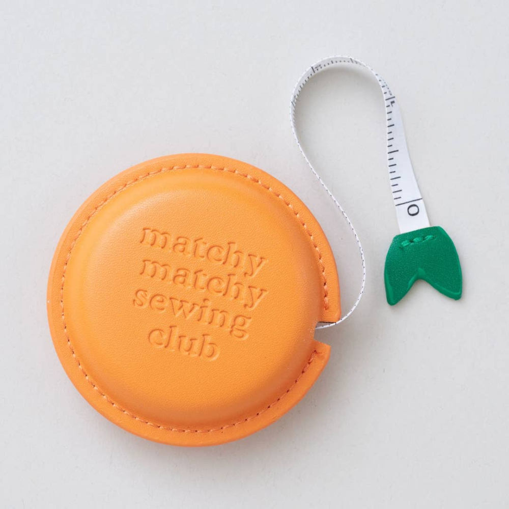 An orange circular leather sewing measuring tape case with the words 'matchy matchy sewing club' embossed on the front, shown on a light gray background with its white measuring tape extended and a green plastic hook.