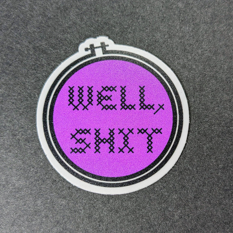 Embroidery hoop sticker with 'WELL SHIT' text on a purple background, placed on a textured gray surface.