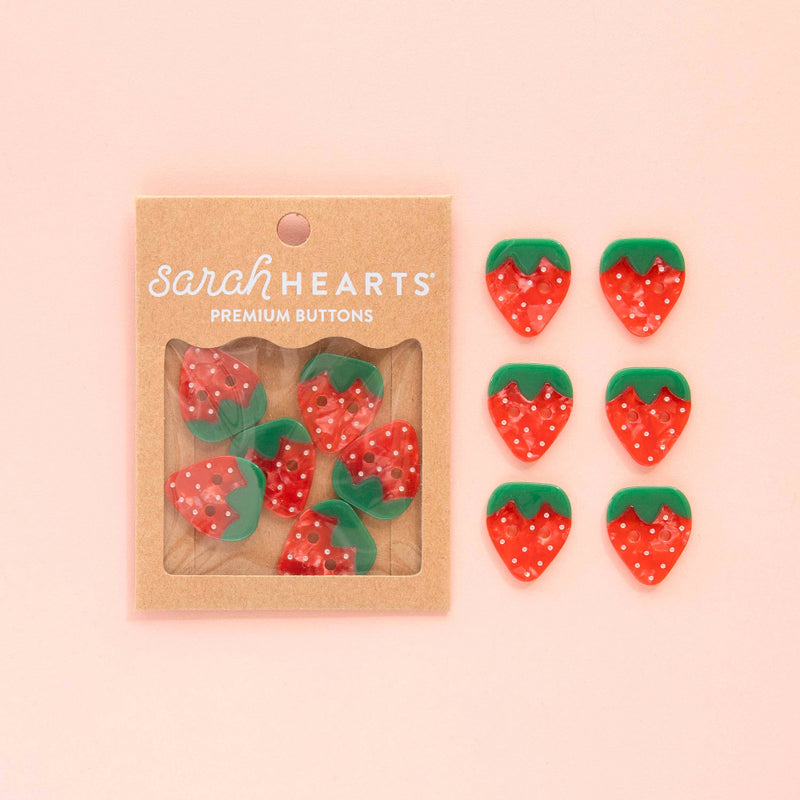 Strawberry-shaped buttons with a 'Sarah Hearts' packaging on a pink background