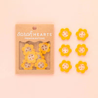 Packaged yellow flower buttons with 'sarah HEARTS' branding on a pink background