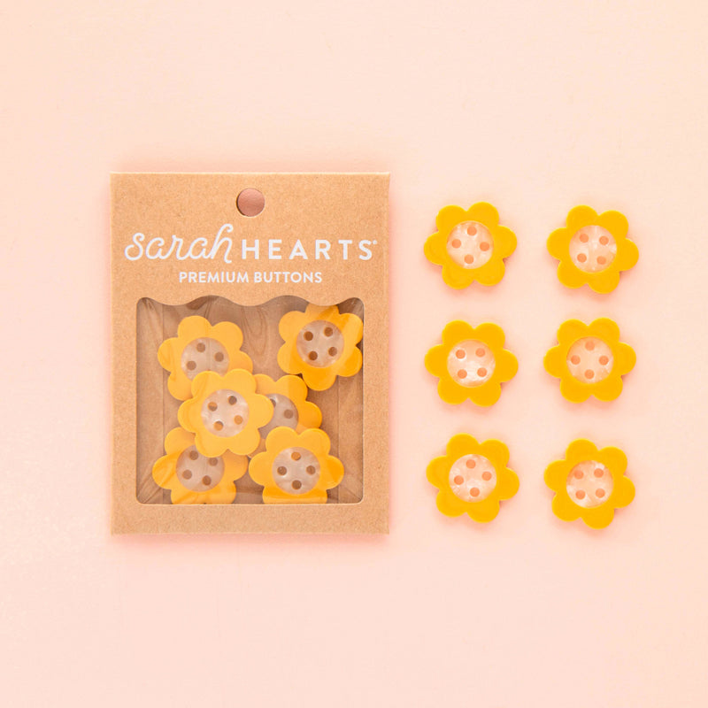 Packaged yellow flower buttons with 'sarah HEARTS' branding on a pink background