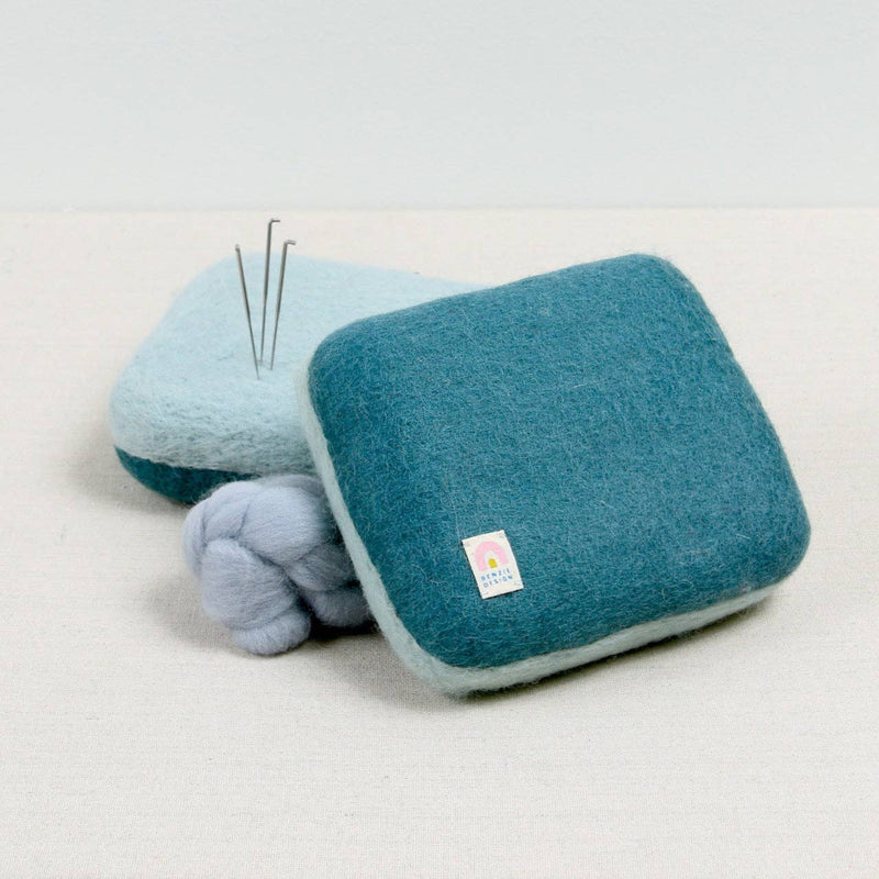 Blue wool needle felting mat with blue fiber and needle felting needles on a white background