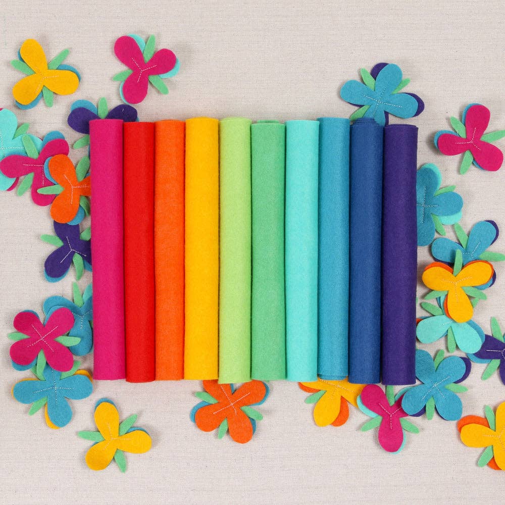 Various shades of felt sheet rolls in shades of a bright rainbow on a textured white background with multicolored felt flowers