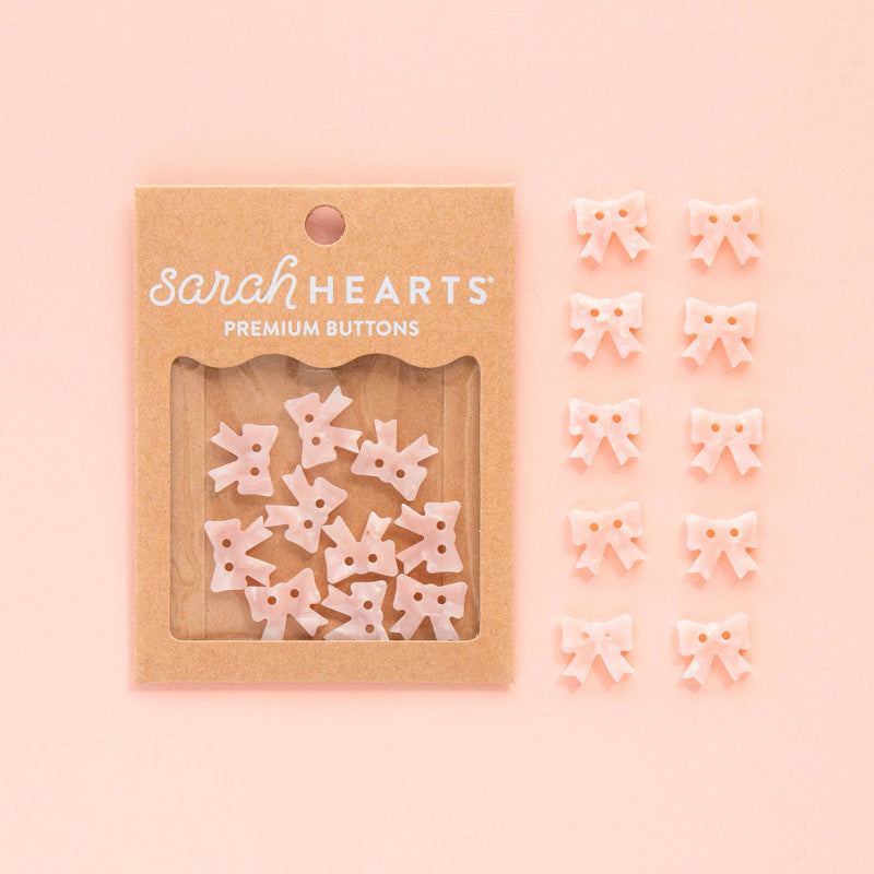 Packaged pink bow buttons with 'sarah HEARTS' branding on a pink background