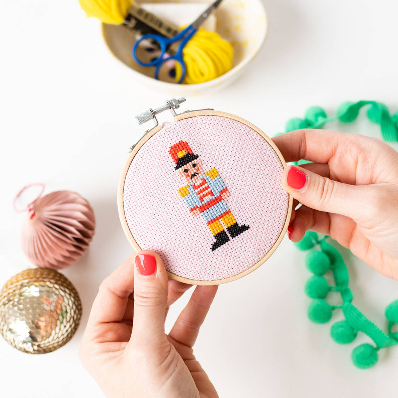 Cross-stitched nutcracker design held in an embroidery hoop with crafting materials in the background.