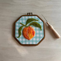 Embroidery hoop with a felted peach on a checkered background, accompanied by a needle and wooden handle.