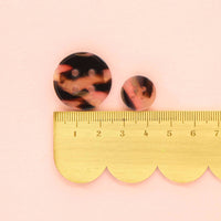 Two tortoiseshell round earrings on a pink background with a gold ruler for scale.
