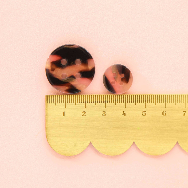 Two tortoiseshell round earrings on a pink background with a gold ruler for scale.